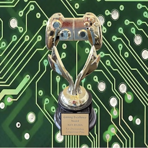 May include: A gold-toned trophy featuring a gaming controller at the top, supported by two curved metallic arms on a black base. A plaque on the base reads: Gaming Excellence Award, ALEX RIVERA, For Outstanding Competitive Play.