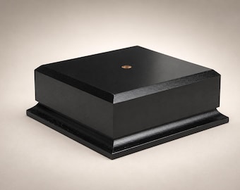 May include: A square black wooden display base with a smooth matte finish and beveled edges. The top surface features a small, centered circular metallic insert. The base has a tiered design with a wider bottom section and a slightly recessed top.
