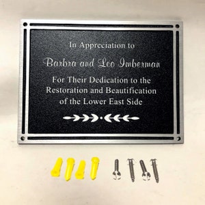 Op de afbeelding: Een zilveren plaquette met zwarte letters die luidt: "In Appreciation to Barbra and Leo Imberman For Their Dedication to the Restoration and Beautification of the Lower East Side".