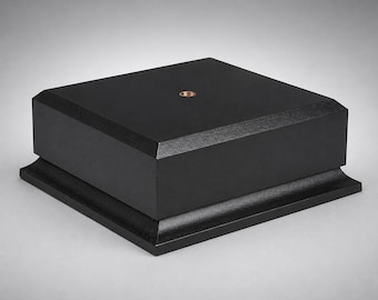 May include: A square black wooden display base with a matte finish and beveled edges. The top surface features a centered circular metal insert with a threaded hole, providing a stable platform for mounting objects or decorative items securely.