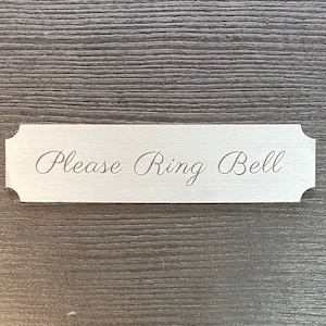 May include: A white rectangular sign with rounded corners and a silver finish. The sign says "Please Ring Bell" in a cursive font.
