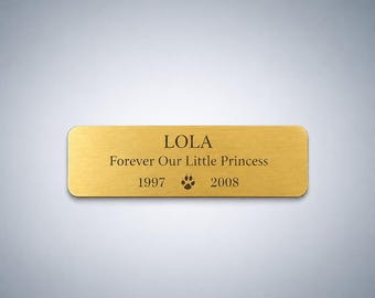 Może przedstawiać: Prostokątna tablica pamiątkowa ze złotego metalu z zaokrąglonymi rogami. Grawerowany tekst brzmi LOLA, Forever Our Little Princess, 1997, a po nim znajduje się mała ikona odcisku łapy oraz rok 2008 wyśrodkowany na szczotkowanej powierzchni metalu.