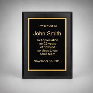 May include: A rectangular black plaque with a gold border and gold text that reads: Presented To John Smith In Appreciation for 25 years of devoted services to our sales team November 15, 2013. The plaque has a smooth, dark frame.