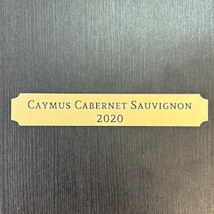 May include: A gold-colored metal plaque with rounded corners and a black inscription that reads "Caymus Cabernet Sauvignon 2020".