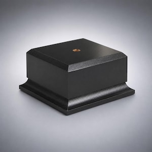 May include: A square black display base with a smooth matte finish and a beveled edge at the bottom. The top surface features a centered circular metallic insert with a threaded hole, providing a stable foundation for mounting various objects.