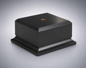 May include: A square black display base with a smooth matte finish and a beveled edge at the bottom. The top surface features a centered circular metallic insert with a threaded hole, providing a stable foundation for mounting various objects.