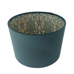 May include: A teal lampshade with a cylindrical shape. The interior has a detailed floral pattern in green, beige, and gold. The lampshade is designed to diffuse light and add a decorative element to any room.