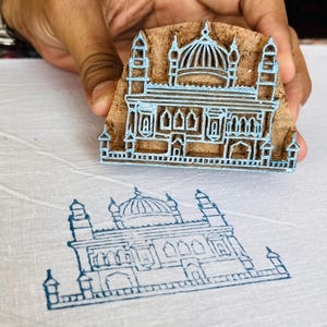 May include: A wooden block stamp with a detailed architectural design in light blue ink. The stamp is held over a white fabric with a matching stamped image. The design features a domed building with intricate details.