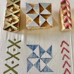 Op de afbeelding: Drie houten blokdrukstempels met geometrische ontwerpen. Eén stempel heeft gouden kruisvormige vormen, een andere zilveren driehoeken en de derde roze chevrons. Bedrukte stof toont bijpassende patronen in groen, blauw en rood.