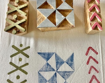 May include: Three wooden block printing stamps with geometric designs. One stamp has gold cross-like shapes, another has silver triangles, and the third has pink chevrons. Printed fabric shows matching patterns in green, blue, and red.