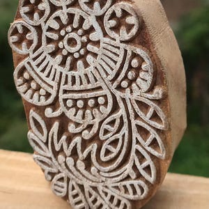 May include: A wooden block printing stamp with a detailed floral design. The stamp features a central circular motif surrounded by intricate patterns of leaves and other shapes. The stamp is made of a light brown wood.