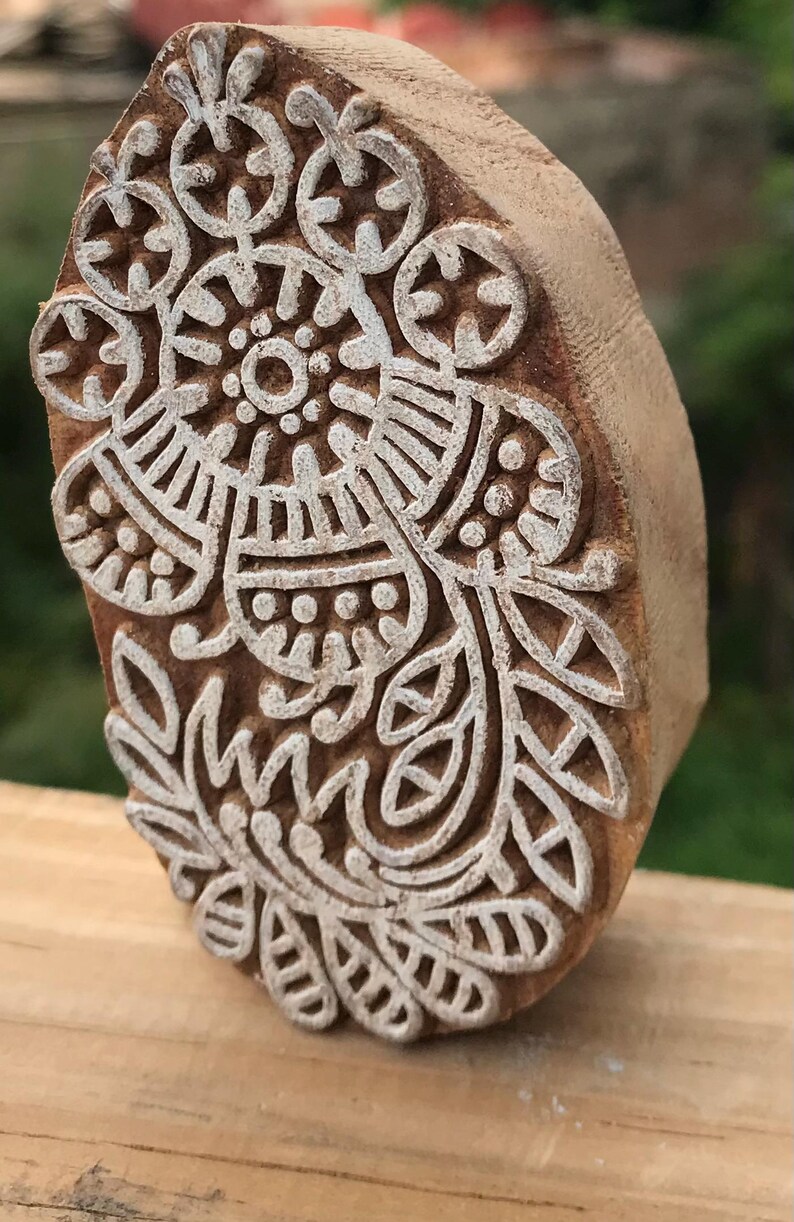May include: A wooden block printing stamp with a detailed floral design. The stamp features a central circular motif surrounded by intricate patterns of leaves and other shapes. The stamp is made of a light brown wood.