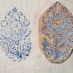 May include: A wooden block with a blue floral design next to a blue floral print on white fabric. The block is oval-shaped and features an intricate pattern of flowers and leaves. The print is a mirror image of the block's design.