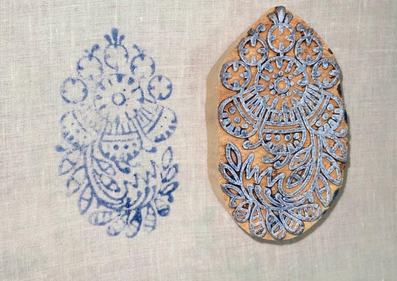 May include: A wooden block with a blue floral design next to a blue floral print on white fabric. The block is oval-shaped and features an intricate pattern of flowers and leaves. The print is a mirror image of the block's design.