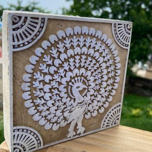 Puede incluir: Bloque de madera cuadrado con un diseño de pavo real tallado. El pavo real es blanco, con una cola detallada en abanico. El fondo es de color marrón claro. Las esquinas del bloque tienen un patrón blanco decorativo.