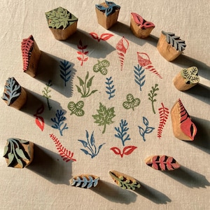 May include: A collection of twelve carved wooden printing blocks arranged in a circle around a central pattern of stamped botanical and seashell motifs in red, green, and blue ink on a light beige fabric surface with visible texture.