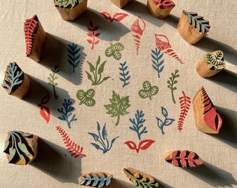 May include: A collection of twelve carved wooden printing blocks arranged in a circle around a central pattern of stamped botanical and seashell motifs in red, green, and blue ink on a light beige fabric surface with visible texture.