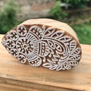 May include: A wooden block with a carved floral design. The block is light brown with a white floral pattern. The design includes a central circular element surrounded by leaves and other decorative shapes. The block is likely used for printing or stamping.