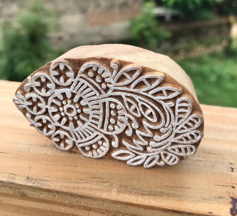 May include: A wooden block with a carved floral design. The block is light brown with a white floral pattern. The design includes a central circular element surrounded by leaves and other decorative shapes. The block is likely used for printing or stamping.