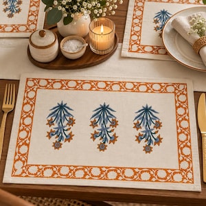 Puede incluir: Un mantel individual rectangular de color blanco roto con un borde floral naranja repetitivo y tres motivos botánicos centrales en azul y naranja. La tela tiene una textura tejida visible y el diseño está impreso en un patrón simétrico.