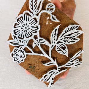 Puede incluir: Un bloque de impresión de madera con un diseño floral blanco. El diseño presenta una flor grande con una flor más pequeña y hojas.