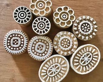May include: A collection of ten wooden printing blocks with various floral and geometric designs. The blocks are light brown with white carved patterns, including circles, flowers, and oval shapes. The blocks are arranged on a light brown wooden surface.