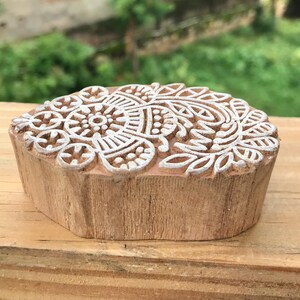 May include: A wooden block with a detailed white floral design on the top surface. The block is a light brown color and has a natural, unfinished appearance. The floral pattern is intricate and symmetrical, suitable for textile printing or crafting.