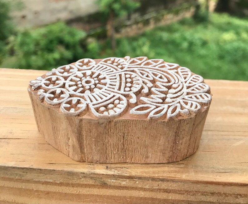 May include: A wooden block with a detailed white floral design on the top surface. The block is a light brown color and has a natural, unfinished appearance. The floral pattern is intricate and symmetrical, suitable for textile printing or crafting.