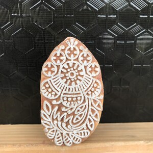 May include: A wooden block with a detailed white floral design. The block is oval-shaped and has a light brown color. The white design includes floral and geometric patterns, suitable for textile printing or crafting.
