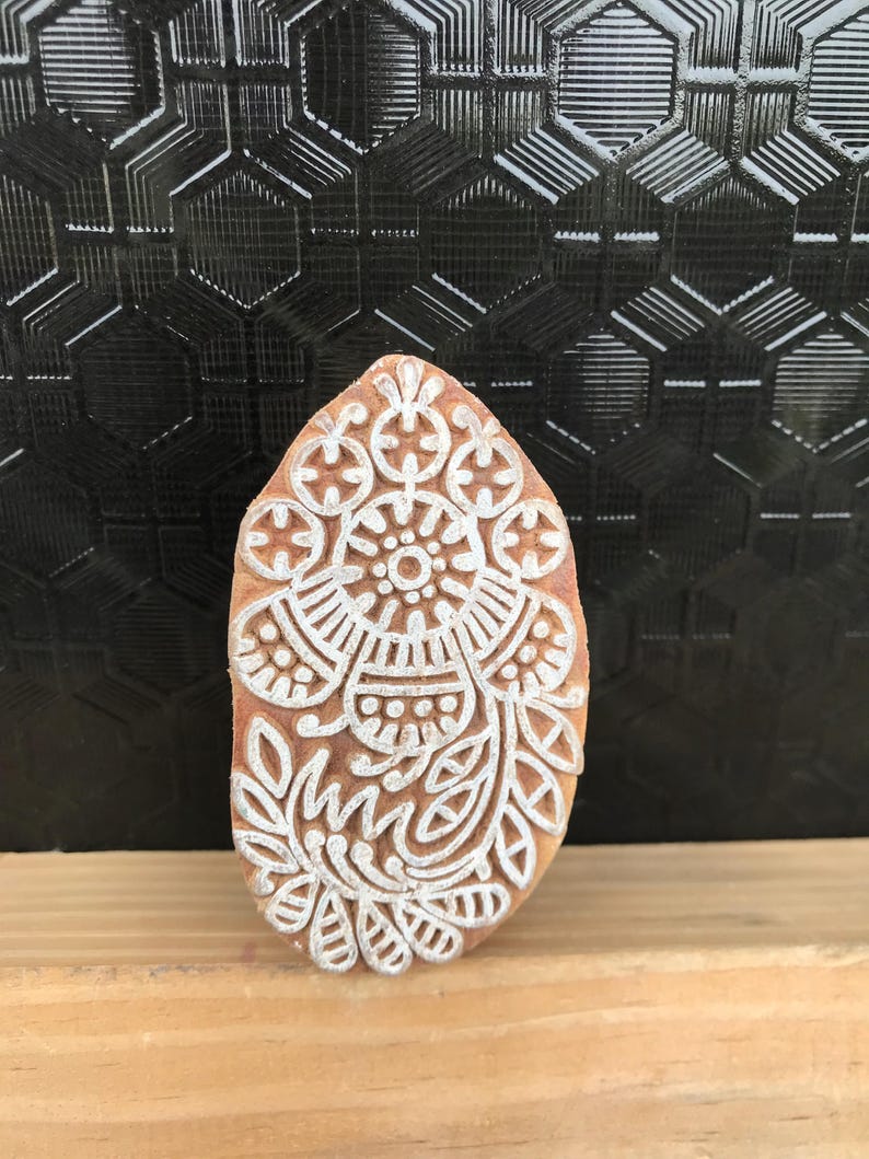 May include: A wooden block with a detailed white floral design. The block is oval-shaped and has a light brown color. The white design includes floral and geometric patterns, suitable for textile printing or crafting.