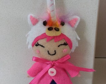 Puede incluir: Una muñeca de fieltro rosa que lleva un disfraz de unicornio blanco con una crin y un cuerno arcoíris. La muñeca tiene un abrigo rosa con un lazo rosa y un botón blanco. La muñeca lleva una falda morada y zapatos negros.