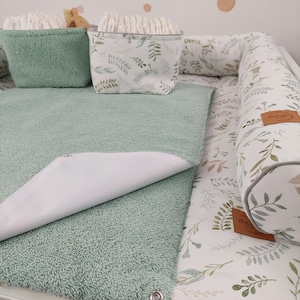Puede incluir: Una alfombra cambiadora verde con un forro impermeable blanco, una cesta de almacenamiento verde con un patr&oacute;n floral blanco y una almohadilla cambiadora blanca y verde con un borde blanco.