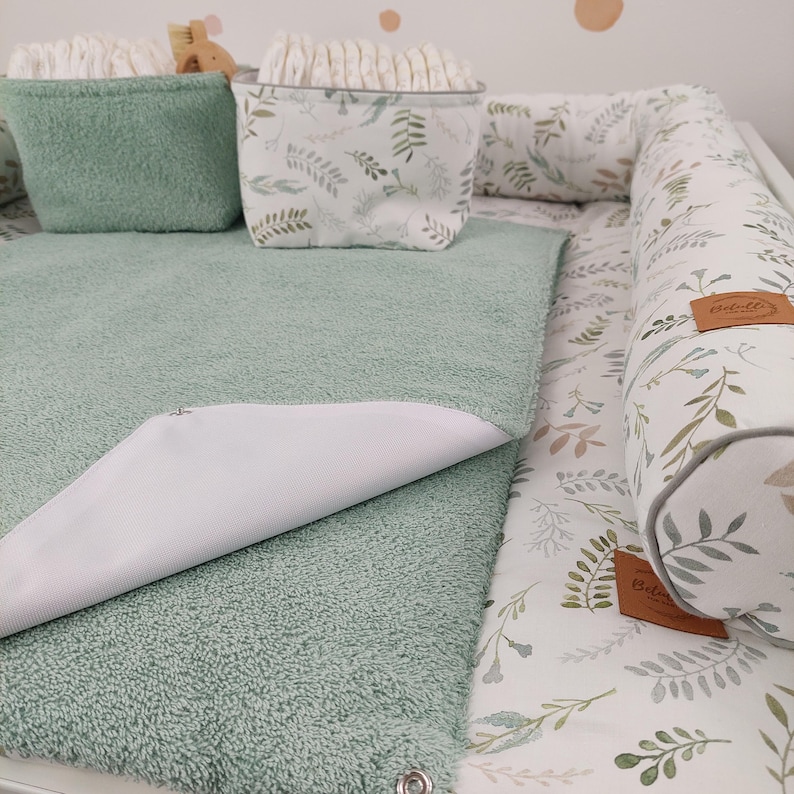 Puede incluir: Una alfombra cambiadora verde con un forro impermeable blanco, una cesta de almacenamiento verde con un patr&oacute;n floral blanco y una almohadilla cambiadora blanca y verde con un borde blanco.