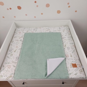 Puede incluir: Una cambiador verde con un forro impermeable blanco. El cambiador est&aacute; encima de una mesa cambiador blanca con un estampado floral. El cambiador est&aacute; hecho de tela suave y felpa.