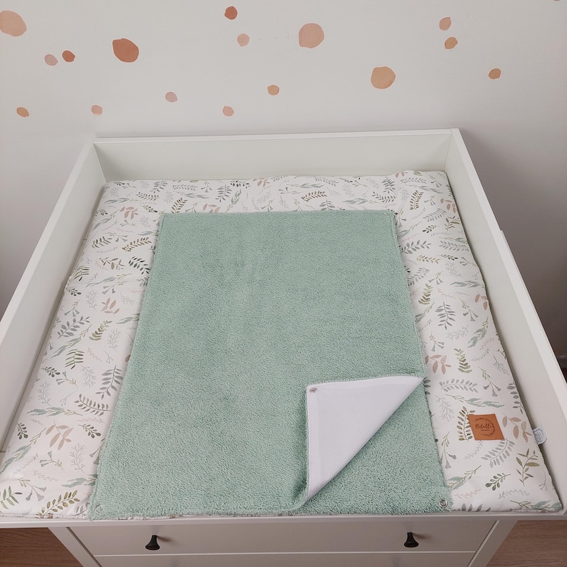 Puede incluir: Una cambiador verde con un forro impermeable blanco. El cambiador est&aacute; encima de una mesa cambiador blanca con un estampado floral. El cambiador est&aacute; hecho de tela suave y felpa.