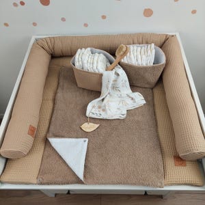 Könnte beinhalten: Eine beige Wickelunterlage mit Waffelstruktur und einem weißen Frottee-Einsatz. Zwei beige Wickelkörbe mit Waffelstruktur, gefüllt mit Windeln und einer Holzbürste, befinden sich auf der Wickelunterlage. Ein weißes Handtuch mit braunem Blumenmuster liegt auf der Wickelunterlage.