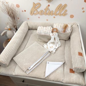 Könnte beinhalten: Eine beige Wickelauflage aus Kord mit erhöhten Seitenpolstern, inklusive zwei passenden Aufbewahrungskörben mit Windeln und einer Holzbürste. Ein gefaltetes weißes Tuch mit braunem Blattmuster und ein Holzring liegen auf der Auflage.