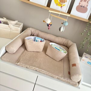 Könnte beinhalten: Eine beige Wickelkommode mit einem Waffelstrick-Stoßfänger und einer Wickelauflage. Zwei passende Körbe liegen auf der Auflage. Darüber zeigen gerahmte Kinderzimmerbilder einen Löwen, einen Bären auf einem Mond und eine Giraffe.