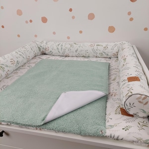 Puede incluir: Una colchoneta cambiadora verde con un forro impermeable, colocada en una mesa cambiadora blanca con un parachoques acolchado de estampado floral blanco y verde. El parachoques tiene una etiqueta de cuero marr&oacute;n con la palabra "Nido" en ella.