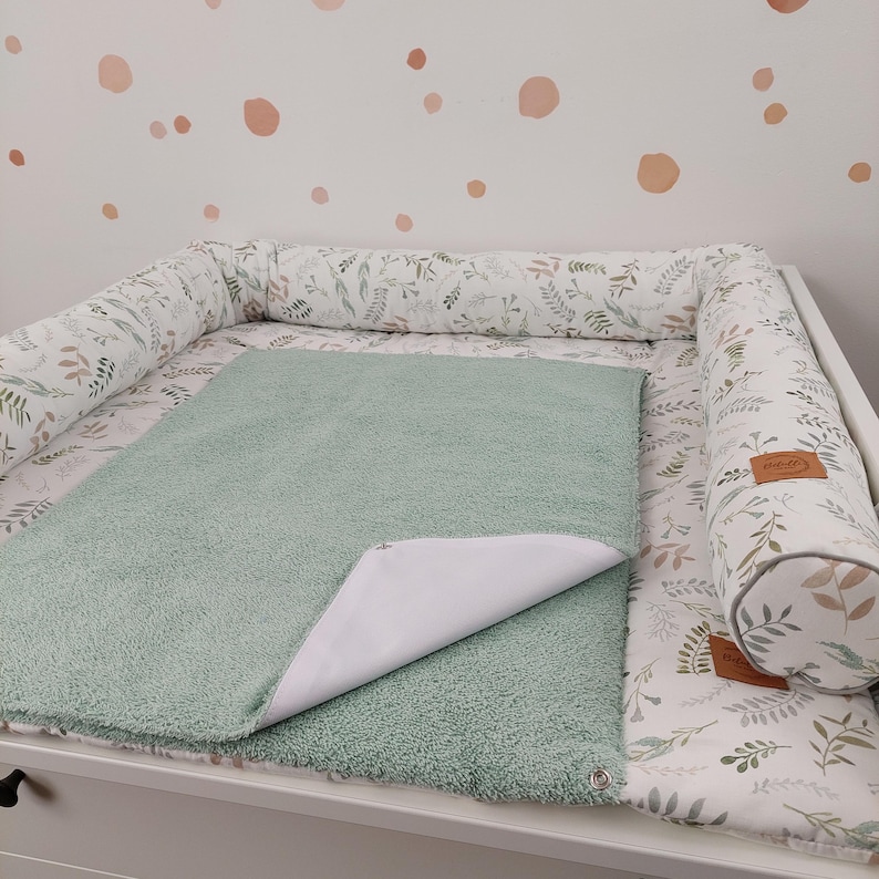 Puede incluir: Una colchoneta cambiadora verde con un forro impermeable, colocada en una mesa cambiadora blanca con un parachoques acolchado de estampado floral blanco y verde. El parachoques tiene una etiqueta de cuero marr&oacute;n con la palabra "Nido" en ella.