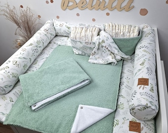 May include: A baby changing pad set featuring a white fabric with a green leaf pattern, including two sage green terry cloth towels, two matching storage baskets filled with nappies, and a long bolster pillow with a brown Betulli brand tag.