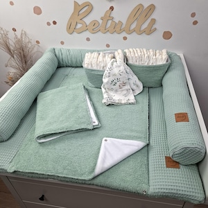 Könnte beinhalten: Ein Wickelunterlagen-Set in Salbeigrün mit Waffelstruktur, bestehend aus zwei passenden Aufbewahrungskörben mit Windeln, einem gefalteten Handtuch und einem Tuch mit Blattmotiven. Das Set enthält eine weiß gefütterte Matte und zwei Seitenrollen.