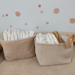 May include: Two beige storage baskets with woven fabric exteriors. One basket is filled with a stack of white cloth nappies, the other with a stack of white cloth nappies and a wooden brush.