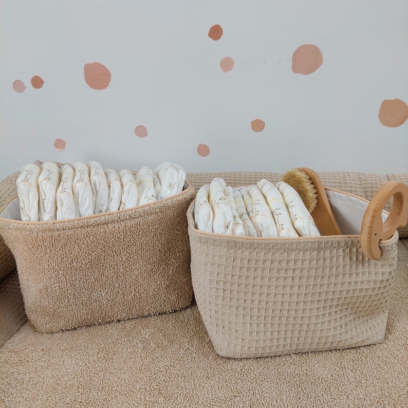 May include: Two beige storage baskets with woven fabric exteriors. One basket is filled with a stack of white cloth nappies, the other with a stack of white cloth nappies and a wooden brush.