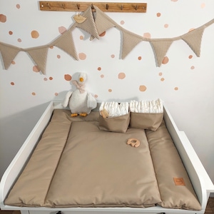 Può includere: Un fasciatoio beige con un fasciatoio abbinato, due contenitori e un anello per la dentizione in legno. Un peluche di oca bianca e una ghirlanda di stoffa beige decorano la parete sopra il tavolo. Il muro ha pois rosa.