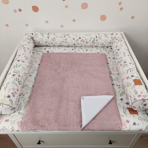 Könnte beinhalten: Ein weißer Wickeltisch mit einem floral gemusterten Stoßfänger und einer rosa Plüsch-Wickelauflage. Der Stoßfänger hat einen weißen Hintergrund mit einem Muster aus rosa, grünen und lila Blumen und Blättern. Die Wickelauflage ist einfarbig, weich rosa.