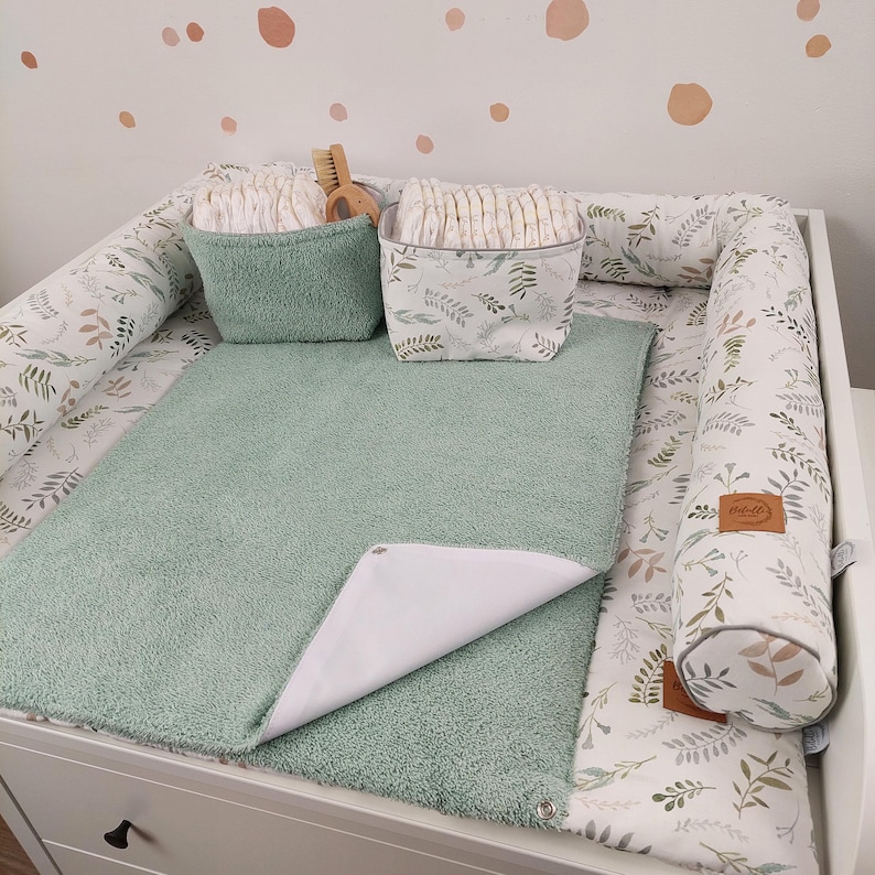 Puede incluir: Una colchoneta cambiadora verde con un forro impermeable blanco, dos parachoques de colchoneta cambiadora con estampado blanco y verde y una cesta blanca con hojas verdes llena de pa&ntilde;ales.