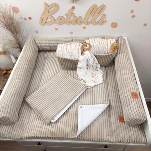 Peut inclure: Un matelas à langer en velours côtelé beige avec des rebords surélevés, comprenant une housse pliée assortie et deux paniers en tissu remplis de couches. Un tissu à motifs blanc et une brosse en bois sont posés entre les paniers.