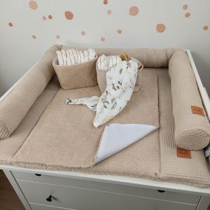 Peut inclure: Une table à langer beige avec un pare-chocs gaufré et un matelas à langer doux. Deux paniers contiennent des couches et une couverture d'emmaillotage à motifs floraux. La table à langer est sur une commode blanche.