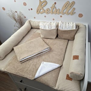 Könnte beinhalten: Ein beiges Wickelunterlagen-Set mit Waffelstruktur, bestehend aus einer weichen Frotteematte mit weißer Unterseite, zwei passenden Aufbewahrungskörben mit Windeln und langen Seitenpolstern. Mit braunen Lederetiketten mit der Aufschrift Betulli.